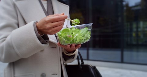 Cinematic close up shot of happy mature asian business woman is eating healthy diet salad from takeaway box while walking in busy city center on way to office during quick lunch break.