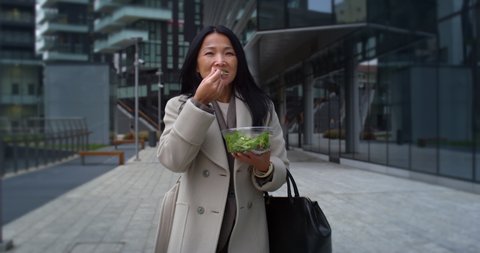 Cinematic shot of happy mature asian business woman is eating healthy diet salad from takeaway box while walking in busy city center on way to office during quick lunch break.