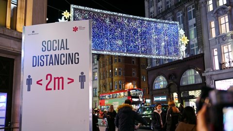 LONDON - NOVEMBER 3, 2020: Covid 19 Social distancing sign and Oxford Street illuminated Christmas lights with Christmas shoppers passing at night