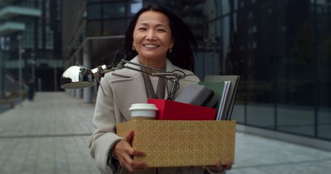 Cinematic close up shot of happy smiling mature asian business woman is holding cardboard box with personal work stuff belongings while walking to new workplace to start her career job.