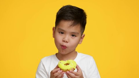 Sweet Tooth. Little Asian Boy Eating Tasty Donut Looking At Camera Standing Over Yellow Studio Background. Kid Enjoying Doughnut And Sugary Food. Unhealthy Junk Food Concept