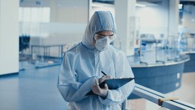 Unrecognizable Male Medical Worker Wearing Protective Hazmat Suit And Respirator Mask Taking Notes Making Record Standing In Modern Clinic Indoors. Slow Motion