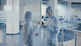 Two Asian Medical Workers Talking Wearing White Hazmat Suits And Protective Respirator Masks Standing In Modern Clinic Or Scientific Center Indoor. Slow Motion
