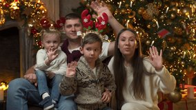 Happy family at home on Christmas Eve enjoy a video call with friends and parents - a young father, mother and two sons look into the camera and wave their hands against the background of the