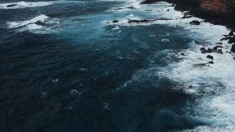 Aerial: coastline of Atlantic Ocean at Tenerife, Canary Islands. Cloudy day with stormy waves. Drone flying over deep blue water. Powerful waves crashing and foaming against black volcanic rocks