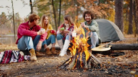 Young friends, men and women, in casual outfit smiling and talking, enjoying tea on picnic, sitting on log in autumn wood near river. Tent, burning campfire and guitar. Slow motion, blurred background