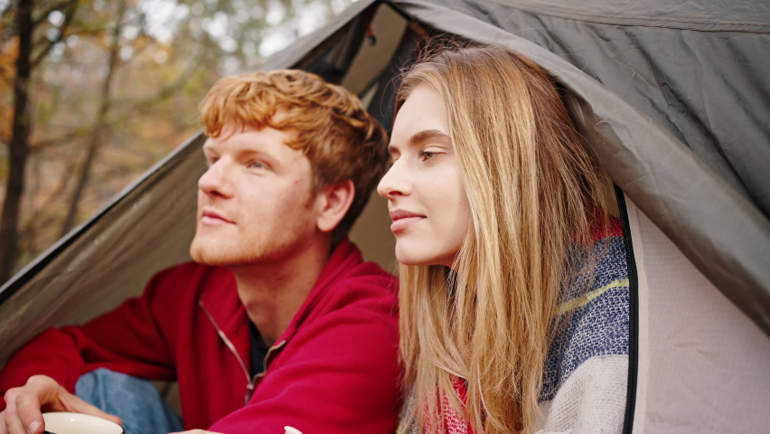 Young woman in checkered plaid is putting her head on shoulder of smiling boyfriend who holding cup of tea while sitting in tent. Autumn day in beautiful forest. Couple in love. Eco tourism. Close up