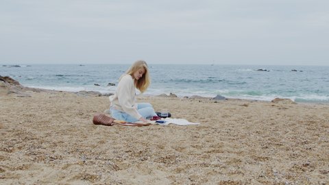 Young woman is sitting on blanket, enjoying her picnic on ocean beach, reading captivating book, renewing after stressful week, Zoom out, Slow motion.