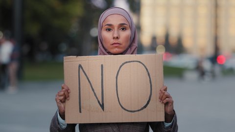 Portrait of muslim scared woman aggressive worried sad stressful girl in hijab stands in city looking at camera cardboard sign banner no protesting denying disagreement, discrimination racism concept