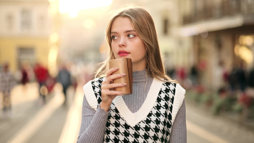 Portrait of joyful caucasian woman smiling and drinking coffee takeaway while walking at city street. Beautiful cheerful young blonde woman walking in the city, holding takeaway coffee cup
