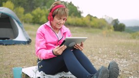 Happy senior woman camping outdoor in the woods while listen to playlist music on tablet computer