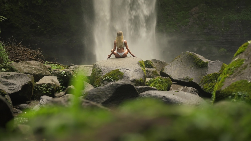 Fit woman in white bikini raising arms in easy pose on rock at powerful waterfall
