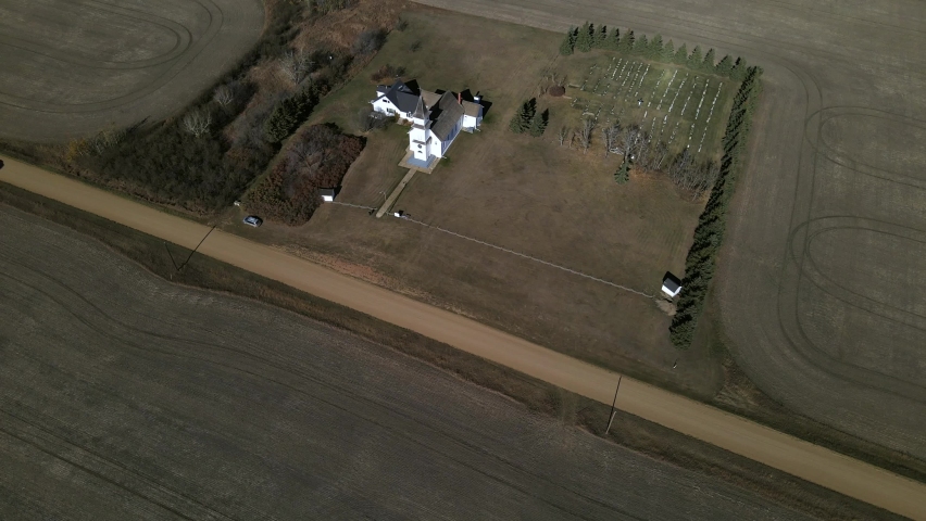 4k aerial footage of St Boniface Church in Flagstaff County, Alberta. Slow push-in movement with 45 degree gimbal angle while car is passing by along a dusty gravel road. Remote tourism concepts.