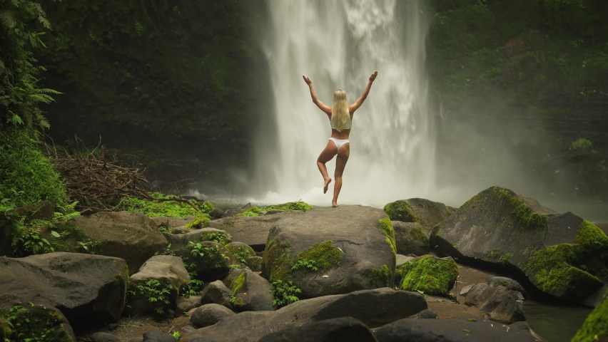 Blond woman in tree yoga pose balance with powerful Nungnung waterfall