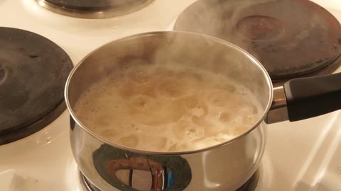 A pot of porridge bubbles on the hob, steam coming off it, in a metal pan with a black handle and 3 other hobs behind it- nearly ready to eat- yum yum! Porridge bubbles on hob, steam coming off