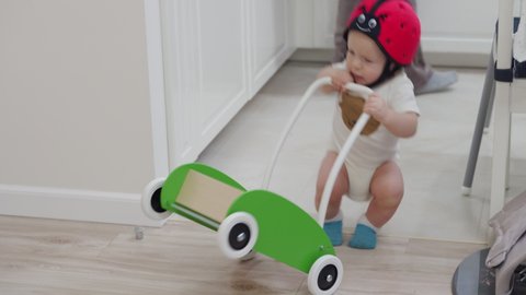 Kid in baby safety helmet learns to walk leaning on push trolley walker, child falling on the floor, head protector helps reduce impact of fall. High quality 4k footage