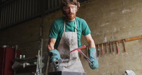 Caucasian male blacksmith wearing safety glasses, hammering hot metal tool on anvil in workshop. small business and craftsmanship at a blacksmiths forge.