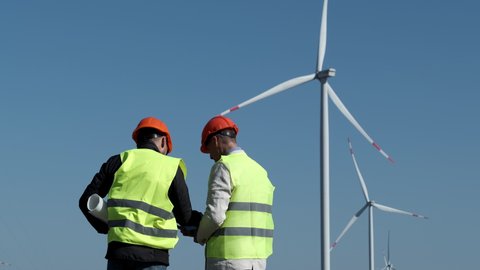 Wind turbines generate energy under blue sky. Professional workers adjust parameters of rotating propellers at offshore station backside view