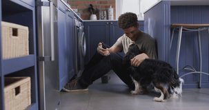 Happy biracial man sitting on floor in kitchen with his pet dog, making video call using smartphone. quality time, relaxing at home.
