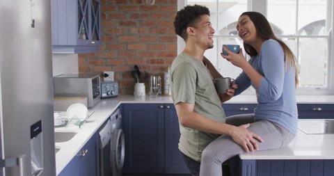 Happy biracial couple sitting on countertop in kitchen, drinking coffee. quality time, relaxing together at home.