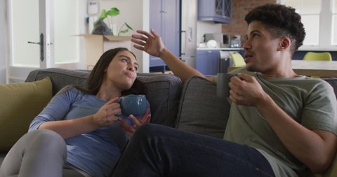 Happy biracial couple sitting on sofa in living room drinking coffee. quality time, relaxing together at home.