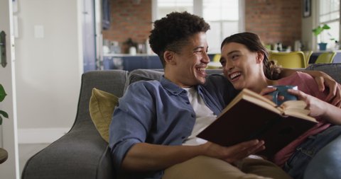 Happy biracial couple sitting on sofa in living room, reading book and drinking coffee. quality time, relaxing together at home.
