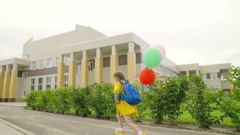 The child runs, studies, holiday, colorful balloons. A kid, a schoolgirl with a backpack, runs to the school building. Happy little girl with a backpack and balloons. Children's school education.