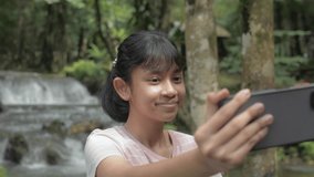 Cheerful Asian pretty girl enjoy taking a selfie video with smartphone near beautiful tropical waterfall background in the jungle. Travel experience. Thailand.