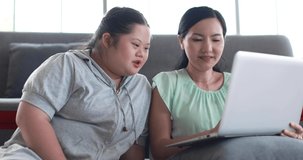 Cheerful loving ethnic mother and girl with down syndrome watching cartoon on netbook together and enjoying weekend at home