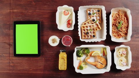 Food delivery service top view, take away meals in disposable containers on wooden table. Close-up of lunch boxes with cooked portions, fish, spaghetti and cakes. Using gadget with chroma green screen