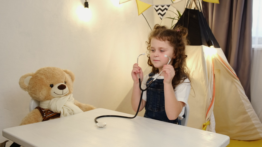Small attractive school cute kid girl pretending to be doctor. Little adorable child using stethoscope, playing as nurse or veterinarian with fluffy toy, treating soft bear sits at table in bedroom Royalty-Free Stock Footage #1082889472