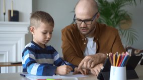 Family education with internet resources. Cute little student draws picture with careful father help during video lesson via contemporary tablet at table spbd
