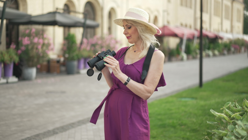Carefree relaxed female tourist standing outdoors in town with binoculars looking at camera smiling. Medium shot side view confident Caucasian woman posing travelling on sunny summer day