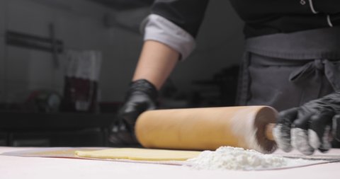 Close-up of a baker's or chef's hand rolling out dough on a white table, with flour so that it doesn't stick to the wooden rolling pin as it rolls out. Close-up of hands in black gloves. Baking