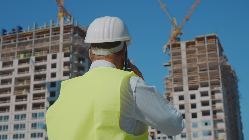 Building inspector wearing hard hat and safety vest looking at skyscrapers under construction and talking on phone, parallax effect. Back view man discussing new city project. Concept of architecture Royalty-Free Stock Footage #1082898730