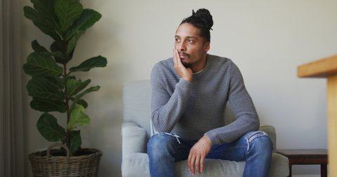 Thoughtful biracial man with dreadlocks in hair bun sitting in armchair in living room looking away. domestic lifestyle, spending time alone at home.