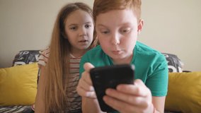 Close up of little smiling brother and sister playing the games on mobile phone at home. Happy small boy and girl watch a funny video on smartphone sitting on sofa at room. Dolly shot Slow motion