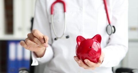 Doctor tosses coin with red pig to piggy bank slow motion 4k movie