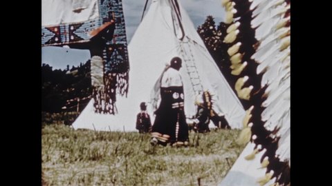 1950s: Woman sits outside of teepee beading clothing. Beaded moccasins and clothing. Man in feather headdress telling story to child.