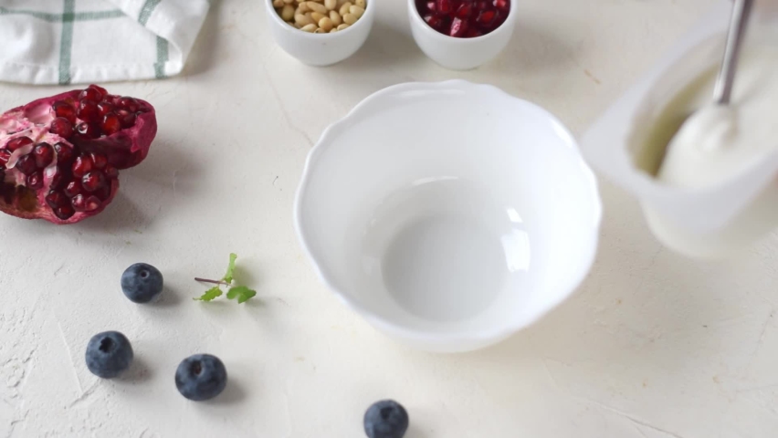 Putting Greek yogurt into the white ceramic bowl with spoon. Thick dairy product and toppings around. Making healthy breakfast