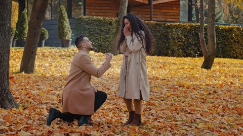 Hispanic loving couple in autumn park man stand in yellow leaves on one knee making marriage proposal girlfriend and puts ring on finger excited happy woman jump and tight hugs sweetheart guy outdoors