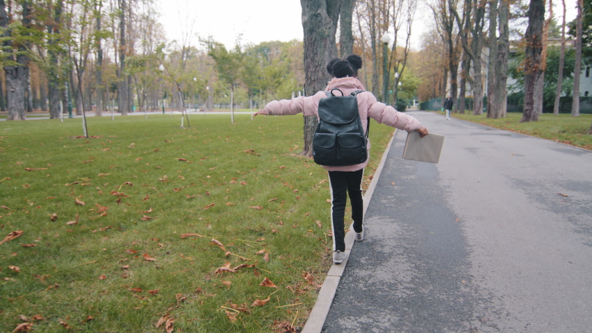 Back view schoolgirl walk on curb little girl with black backpack keep balance child going to school holding book in hand unknown kid walking in city park after study african american pupil play alone Royalty-Free Stock Footage #1082913793