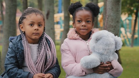 Girls looking at camera two friends sitting on bench in park kid gives teddy bear little sisters play outdoors schoolgirls playing together after school african american children smile happy childhood