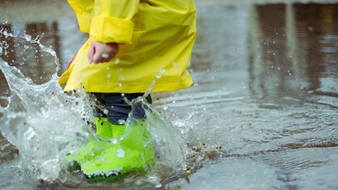 child in green rubber boots and yellow raincoat is jumping in puddle in the rain. child in rubber boots jumping in a puddle. slow motion video 