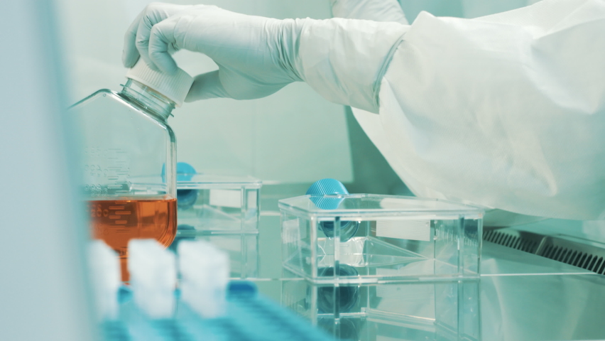 Handheld shot of a Caucasian female lab scientist in a full protective suit, with glasses, face mask, and gloves, taking a liquid with a pipette and putting it in a cell culture flask. Royalty-Free Stock Footage #1082916694