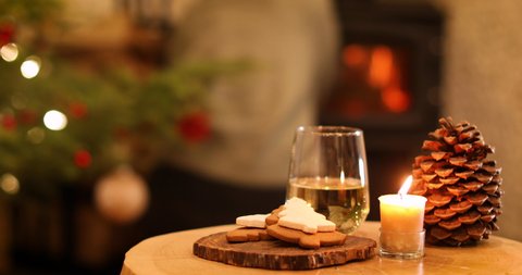 Cookies, glass of white wine, candlelight	and pine cone on rustic table with woman adding firewood to a fire in background.