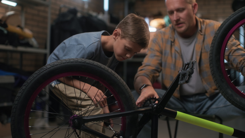 Repair bicycle by small boy in garage. Friendly father helps son to fix bike. Maintenance care and installation of bicycle. Happy dad explains to young cycle repairman indoors. Fatherhood and grow up Royalty-Free Stock Footage #1082924749