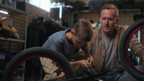 Repair bicycle by small boy in garage. Friendly father helps son to fix bike. Maintenance care and installation of bicycle. Happy dad explains to young cycle repairman indoors. Fatherhood and grow up