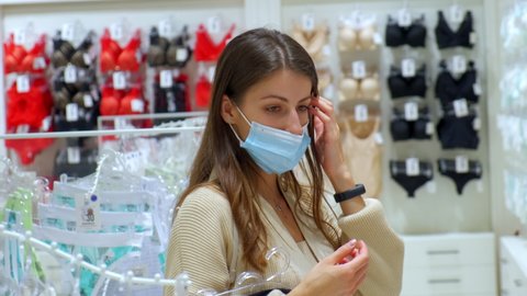 Shopping at quarantine lockdown. Pretty lady with disposable mask holds hangers with bras choosing lingerie in fashion store
