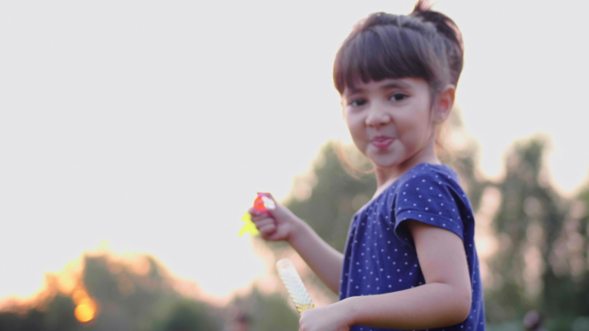 A close-up beautiful happy shot of a cute and adorable little girl child is blowing soap water bubbles against the sun in the park or garden. 